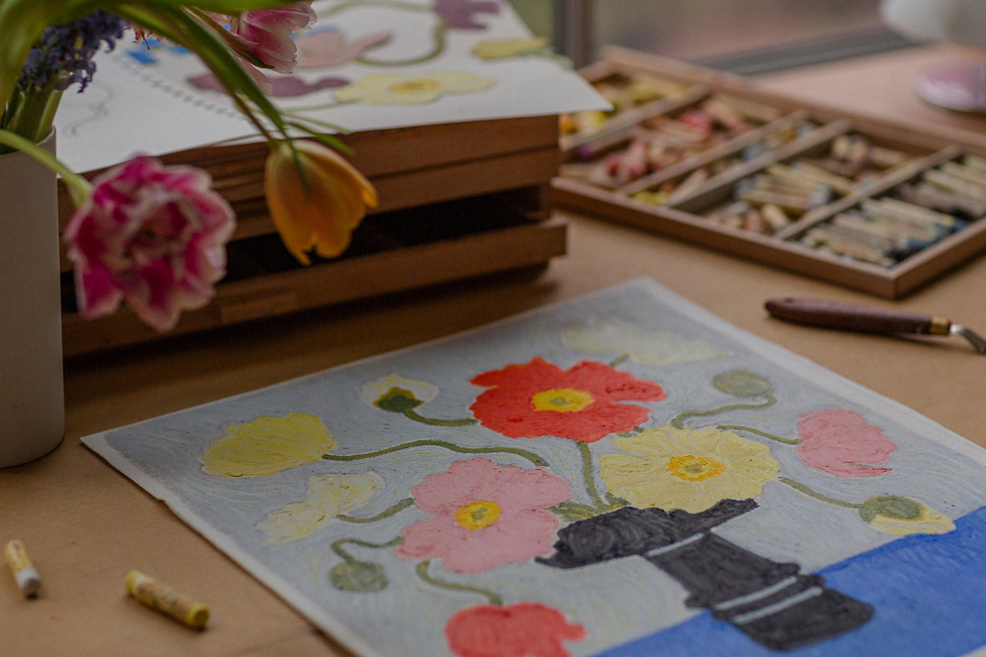A finished oil pastel painting is sitting on a desk, surrounded by a wooden tray of oil pastels and a bouquet of flowers.