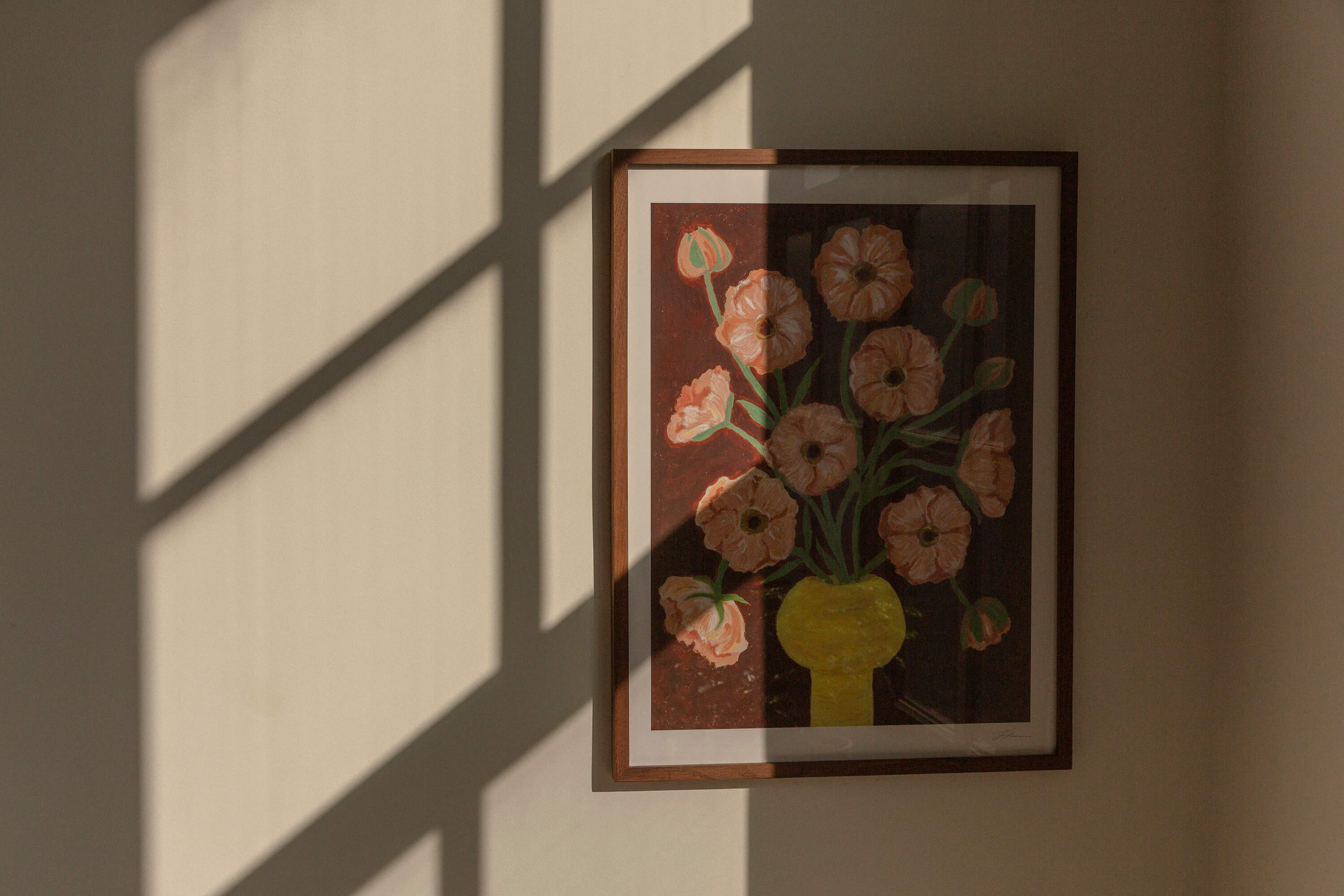 Framed artwork of a floral bouquet of pink butterfly ranunculus in a yellow vase and brown background. The artwork is hung up on a wall with window shadows.