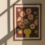 Framed artwork of a floral bouquet of pink butterfly ranunculus in a yellow vase and brown background. The artwork is hung up on a wall with window shadows.