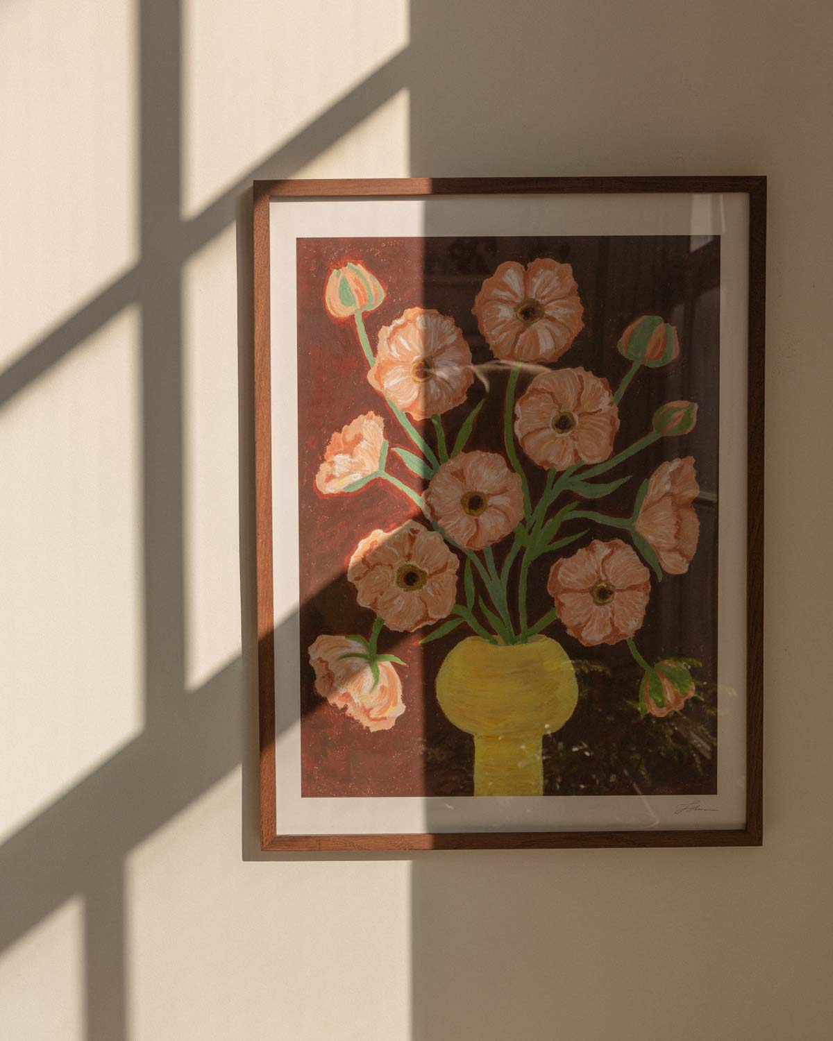 Framed artwork of a floral bouquet of pink butterfly ranunculus in a yellow vase and brown background. The artwork is hung up on a wall with window shadows.