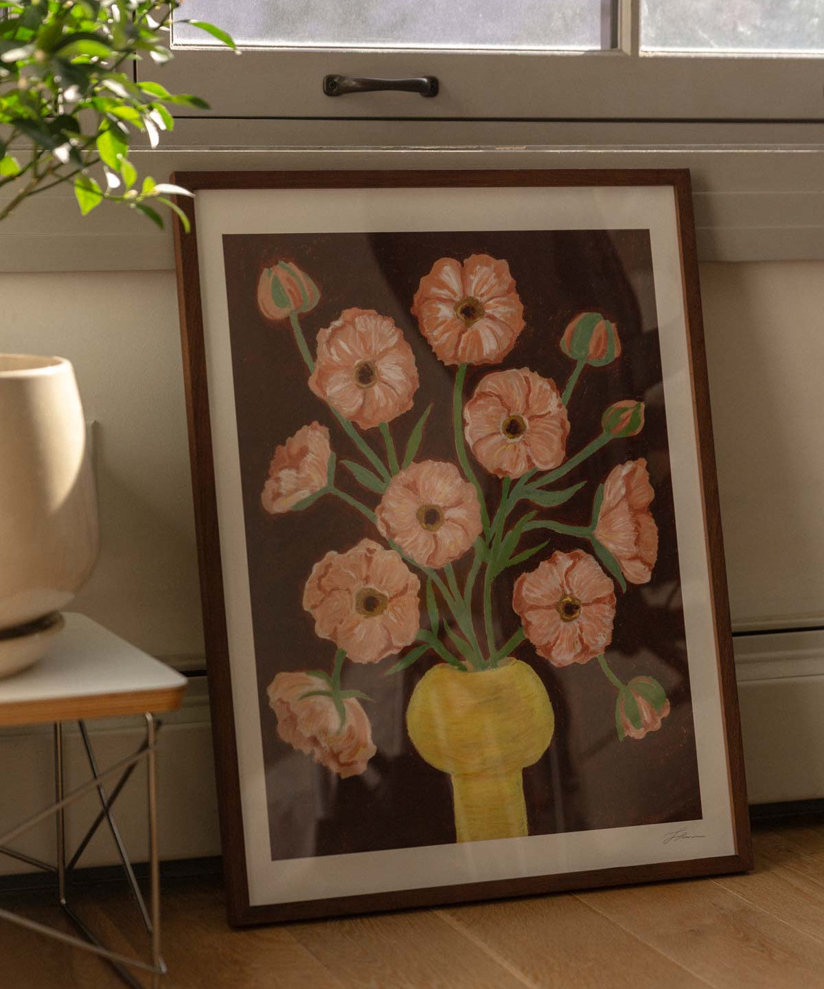 Framed artwork of a floral bouquet of pink butterfly ranunculus in a yellow vase and brown background. The artwork is leaning against the wall with a window, on a wooden floor next to a houseplant. 