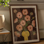 Framed artwork of a floral bouquet of pink butterfly ranunculus in a yellow vase and brown background. The artwork is leaning against the wall with a window, on a wooden floor next to a houseplant. 