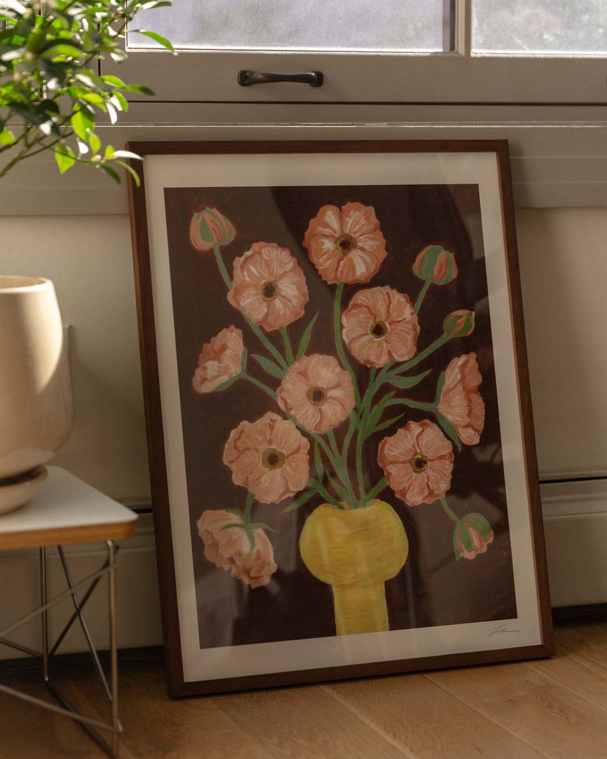 Framed artwork of a floral bouquet of pink butterfly ranunculus in a yellow vase and brown background. The artwork is leaning against the wall with a window, on a wooden floor next to a houseplant. 