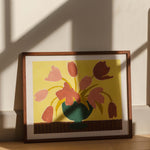 Framed artwork of pink tulips in a green vase on a butter yellow background, placed on a wooden surface with sunlight casting shadows.