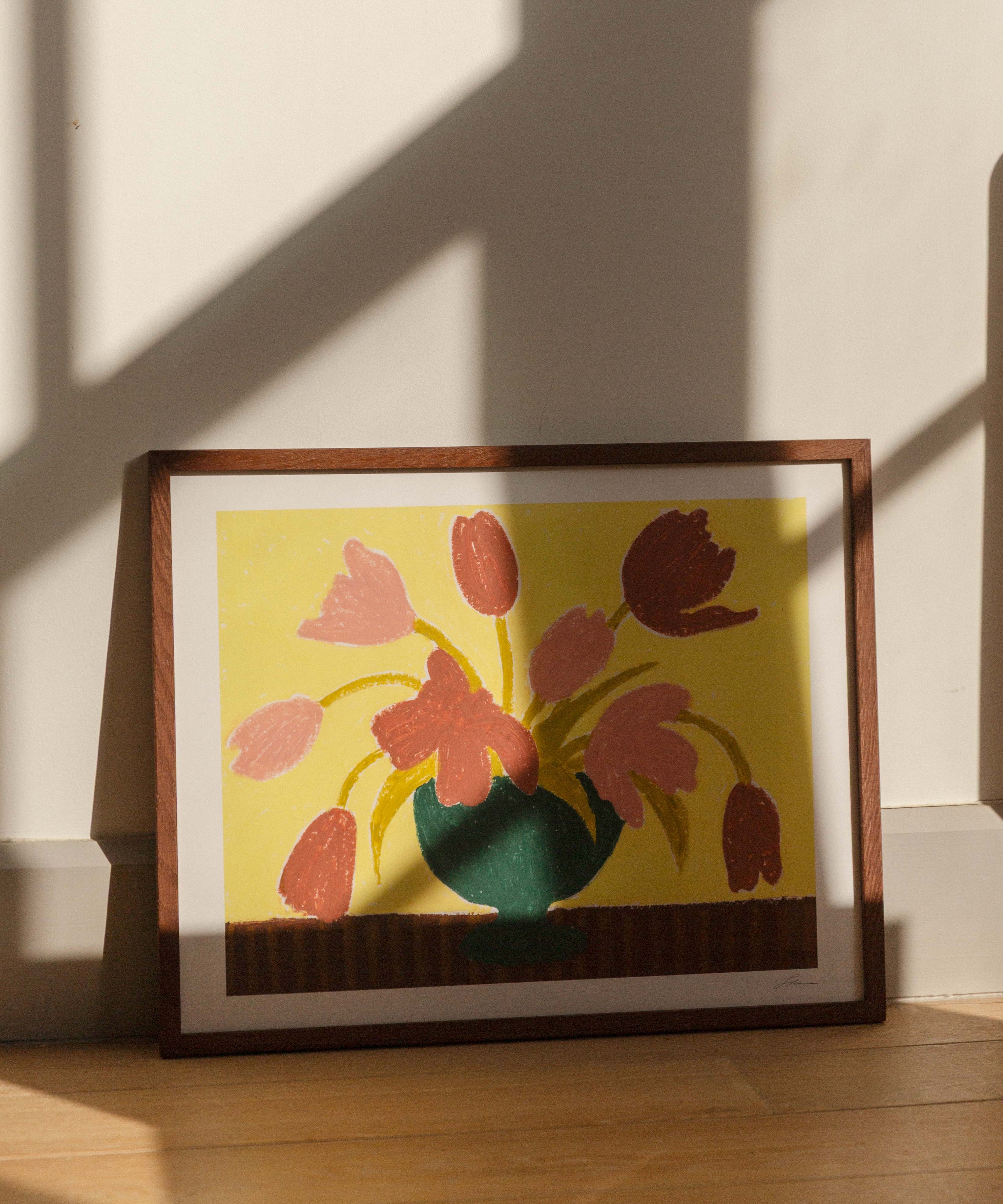 Framed artwork of pink tulips in a green vase on a butter yellow background, placed on a wooden surface with sunlight casting shadows.