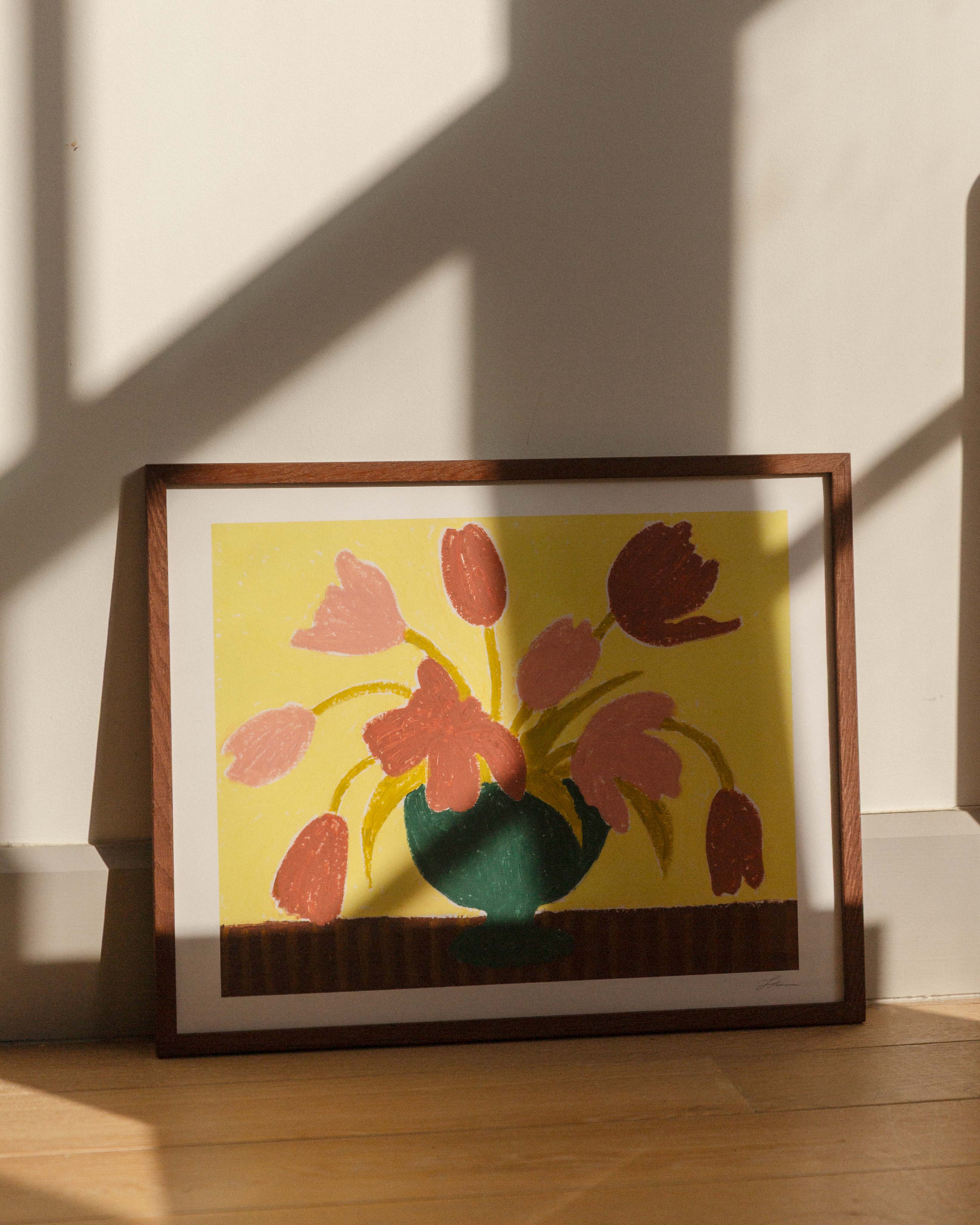 Framed artwork of pink tulips in a green vase on a butter yellow background, placed on a wooden surface with sunlight casting shadows.