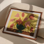 Framed artwork of pink tulips in a green vase on a butter yellow background, resting on top of a textured ivory armchair with hard window sunlight shadows. 