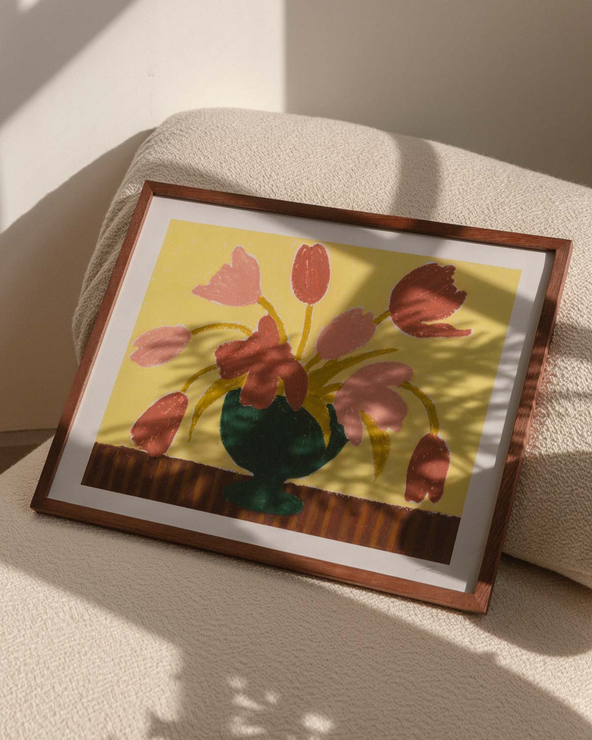 Framed artwork of pink tulips in a green vase on a butter yellow background, resting on top of a textured ivory armchair with hard window sunlight shadows. 