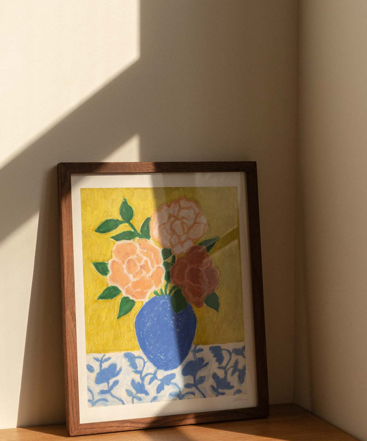 Framed artwork of a blue vase with 3 large roses in varying shades of pink sitting on top of a floral patterned tablecloth against a textured yellow green background. The artwork is sitting on a wooden table, with hard window sunlight shadows.