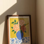 Framed artwork of a blue vase with 3 large roses in varying shades of pink sitting on top of a floral patterned tablecloth against a textured yellow green background. The artwork is sitting on a wooden table, with hard window sunlight shadows.