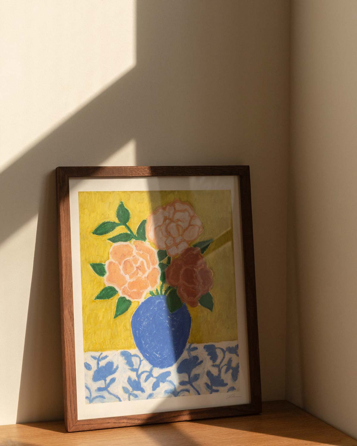 Framed artwork of a blue vase with 3 large roses in varying shades of pink sitting on top of a floral patterned tablecloth against a textured yellow green background. The artwork is sitting on a wooden table, with hard window sunlight shadows.