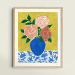 Framed artwork of a blue vase with 3 large roses in varying shades of pink sitting on top of a floral patterned tablecloth against a textured yellow green background.