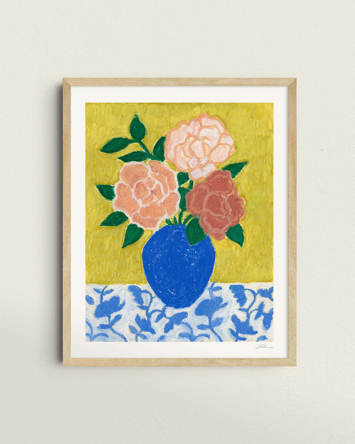 Framed artwork of a blue vase with 3 large roses in varying shades of pink sitting on top of a floral patterned tablecloth against a textured yellow green background.